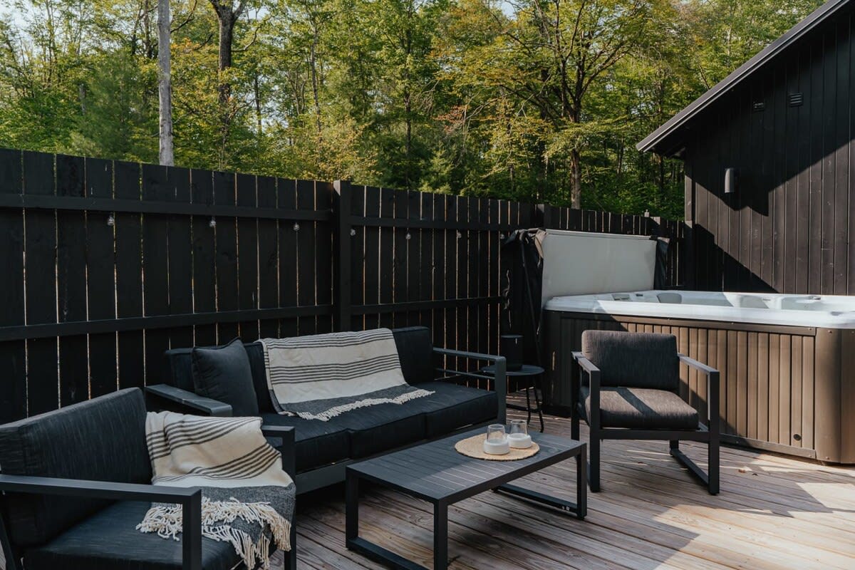 Outdoor lounge area on a wooden deck with black modern patio furniture, including a sofa and chairs, next to a hot tub and a black privacy fence.
