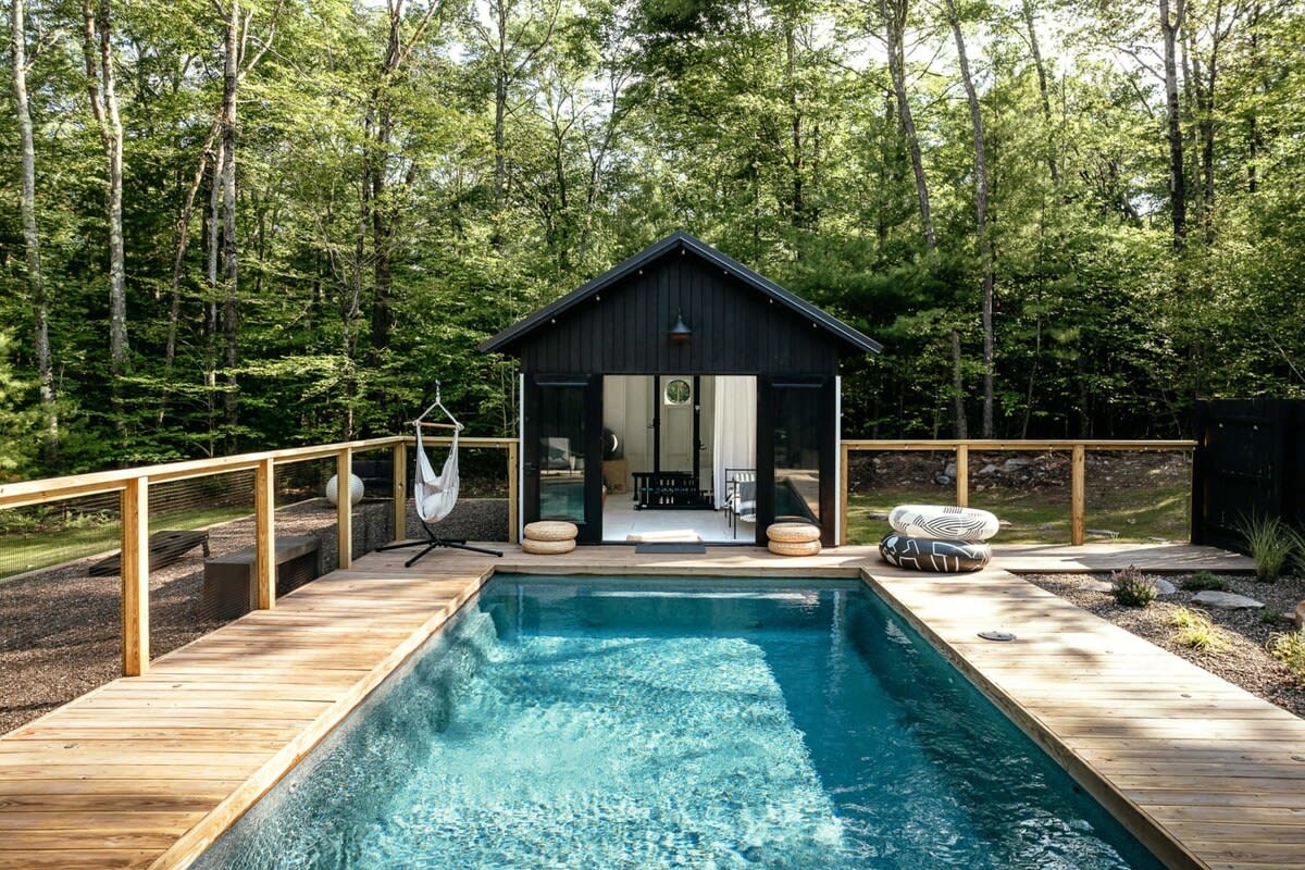 A rectangular swimming pool with a wooden deck is set in a clearing, bordered by a black cabin with large glass doors and a dense green forest. A hammock chair hangs near the cabin.