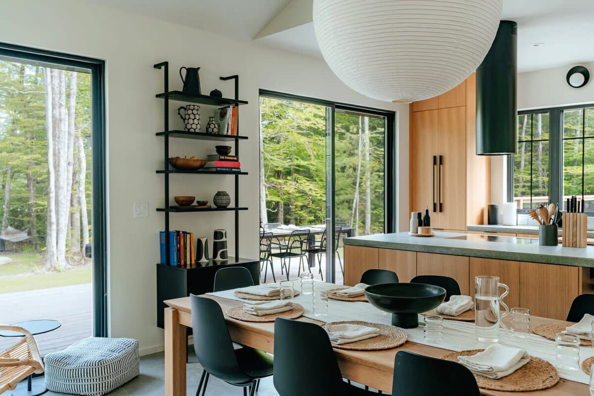 A stylish dining area featuring a light wood table set with black chairs and woven placemats, illuminated by a large white spherical pendant light, adjacent to a black shelving unit and large sliding glass doors.