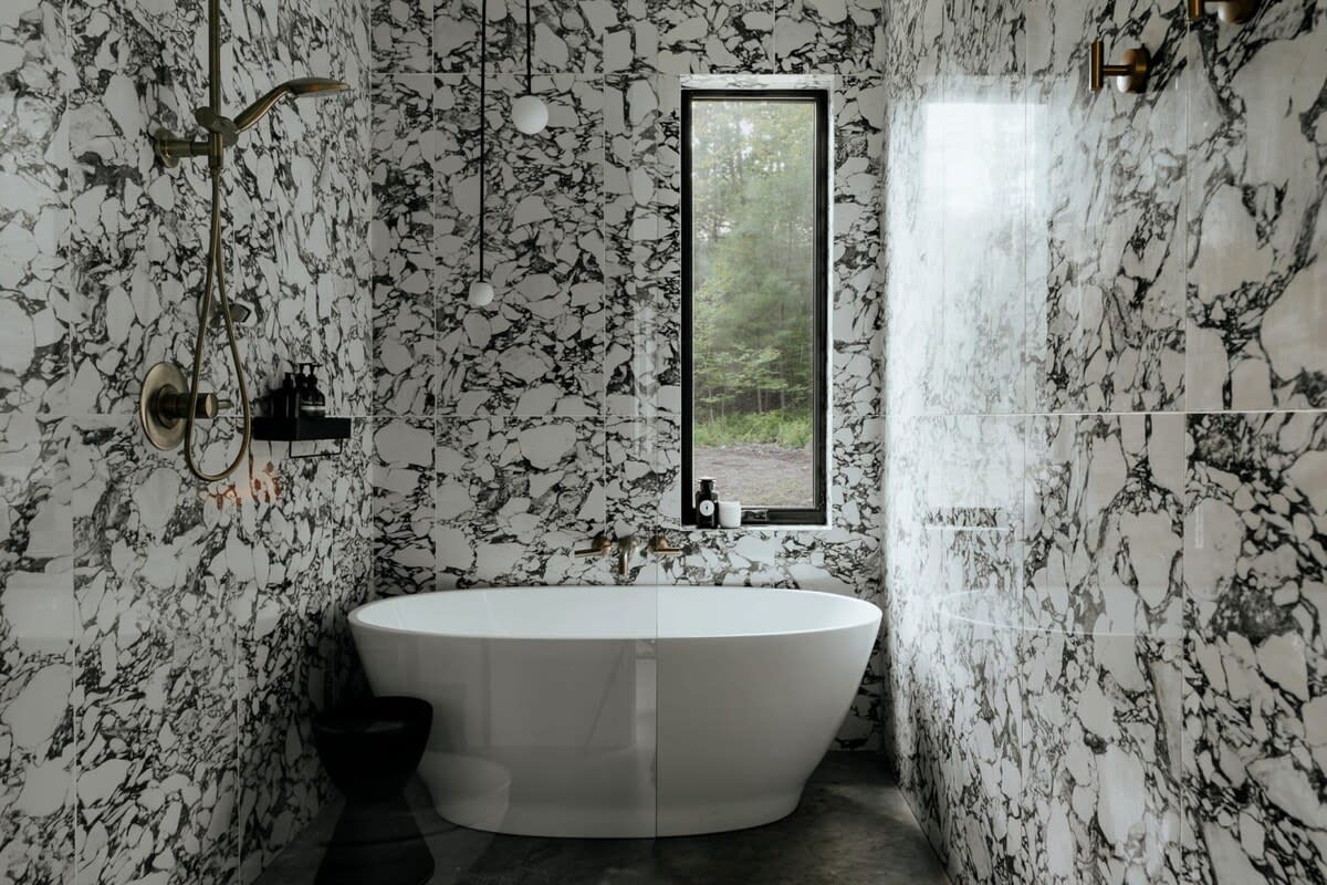 Luxurious bathroom featuring a freestanding white bathtub, a walk-in shower with a rain showerhead, and walls covered in dramatic black and white marble-patterned tiles. A tall window offers a glimpse of the forest.