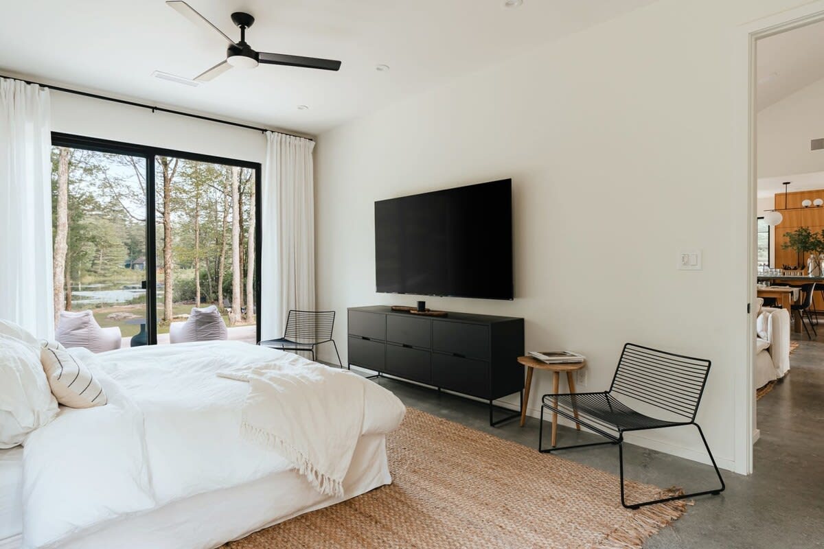 Bright bedroom with a white bed, black metal chairs, a black media console with a large TV, and a sliding glass door leading to an outdoor area with lake views.