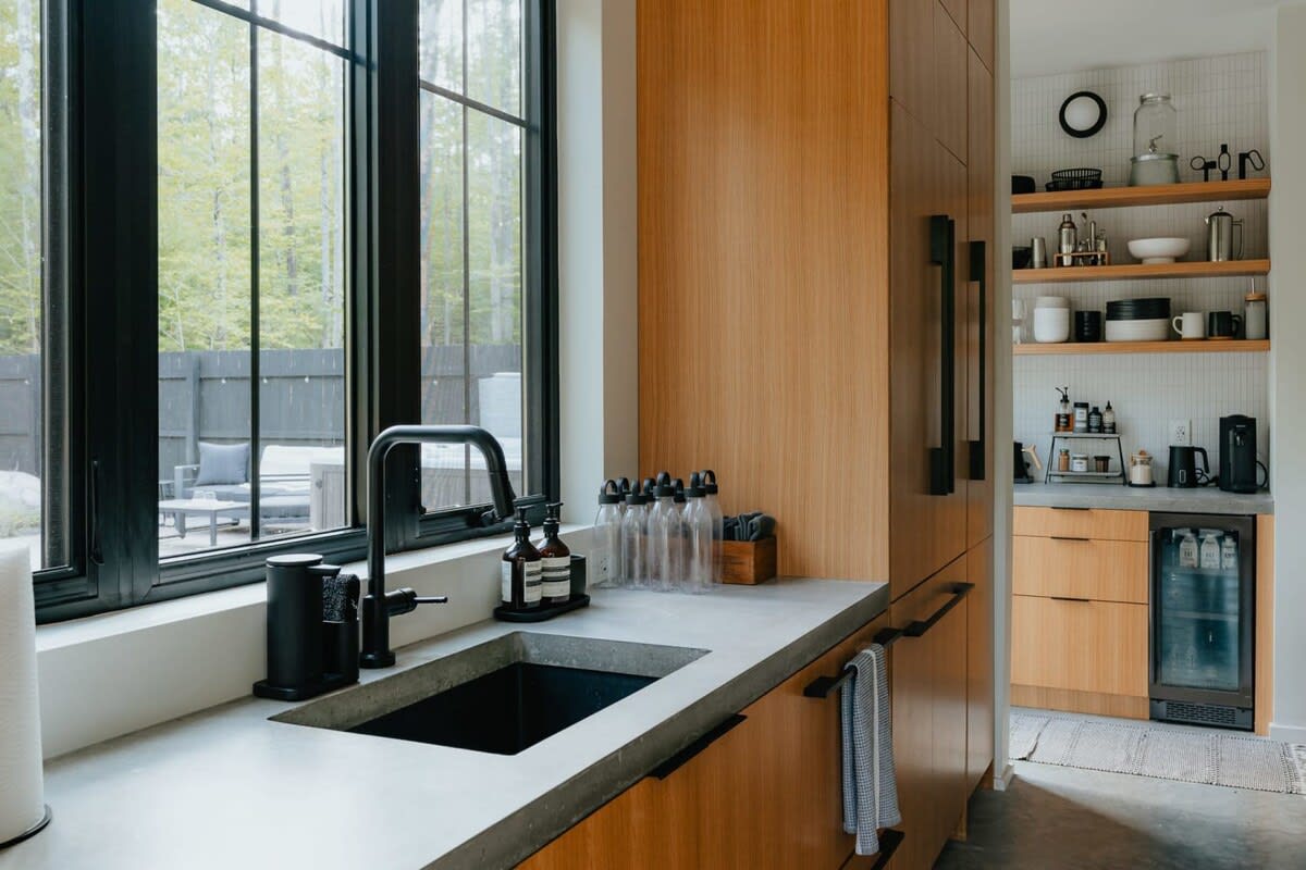 Modern kitchen with light wood cabinetry, concrete countertops, a black sink, and a large black-framed window overlooking a wooded backyard with outdoor seating.