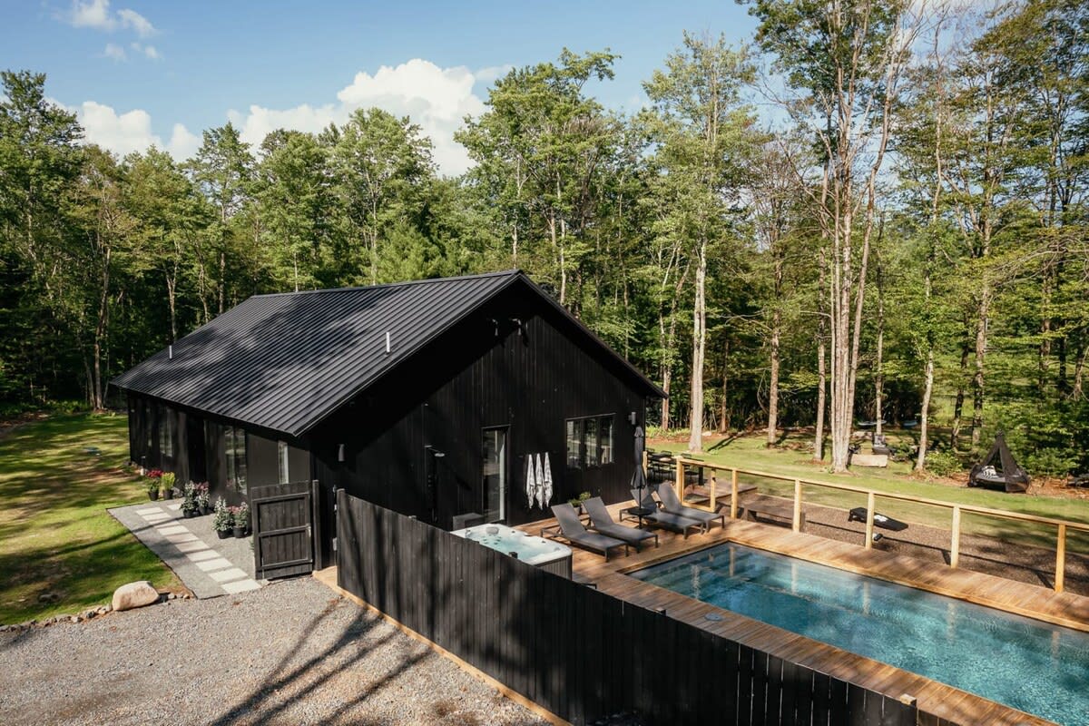 A modern black cabin with a dark metal roof, surrounded by lush green trees. In the foreground, there's a rectangular swimming pool with a wooden deck, a hot tub, and lounge chairs, all enclosed by a dark wooden fence.