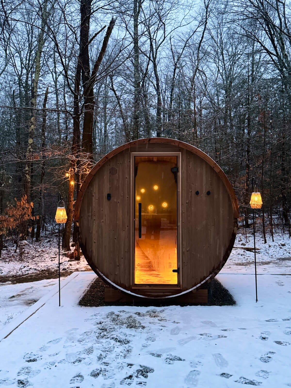 A barrel-shaped wooden sauna with a glass door is nestled in a snowy, wooded area, illuminated by warm interior lights and two outdoor lanterns, creating a cozy winter scene.