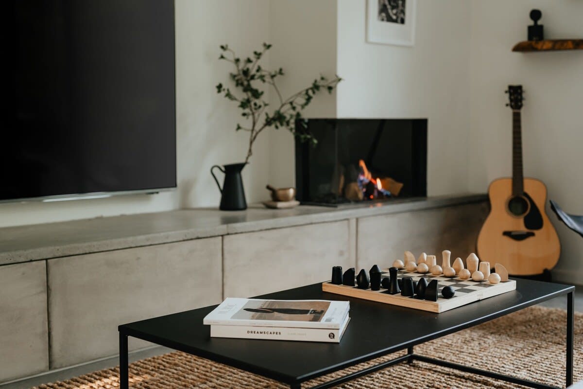 A close-up of the living room's modern black coffee table, displaying a wooden chess set and books, with a contemporary fireplace and an acoustic guitar visible in the background.