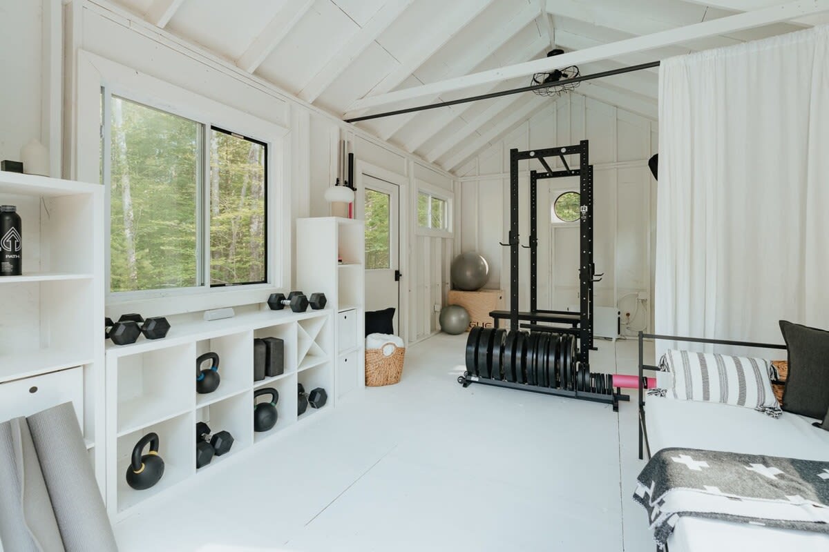 A bright, all-white shed-like structure converted into a home gym, featuring exposed ceiling beams, large windows overlooking a forest, and various weights, a squat rack, and exercise balls. A white daybed with pillows is also visible.