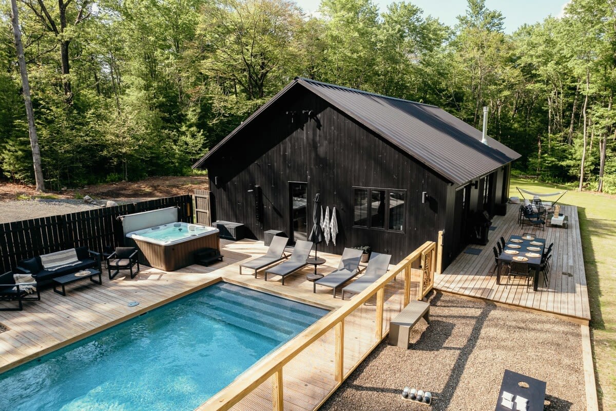 Aerial view of the black-sided cabin with a large wooden deck, featuring an outdoor swimming pool, a hot tub, and various seating areas, surrounded by a dense forest.
