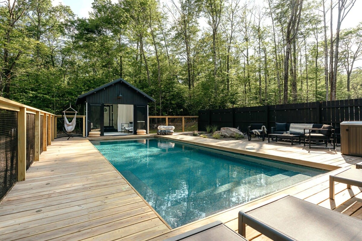 A rectangular swimming pool with a light blue interior, surrounded by a light-colored wooden deck and a wooden fence. Beyond the pool, a small black cabin with an open door reveals a hammock inside, and an outdoor seating area with dark furniture is visible.