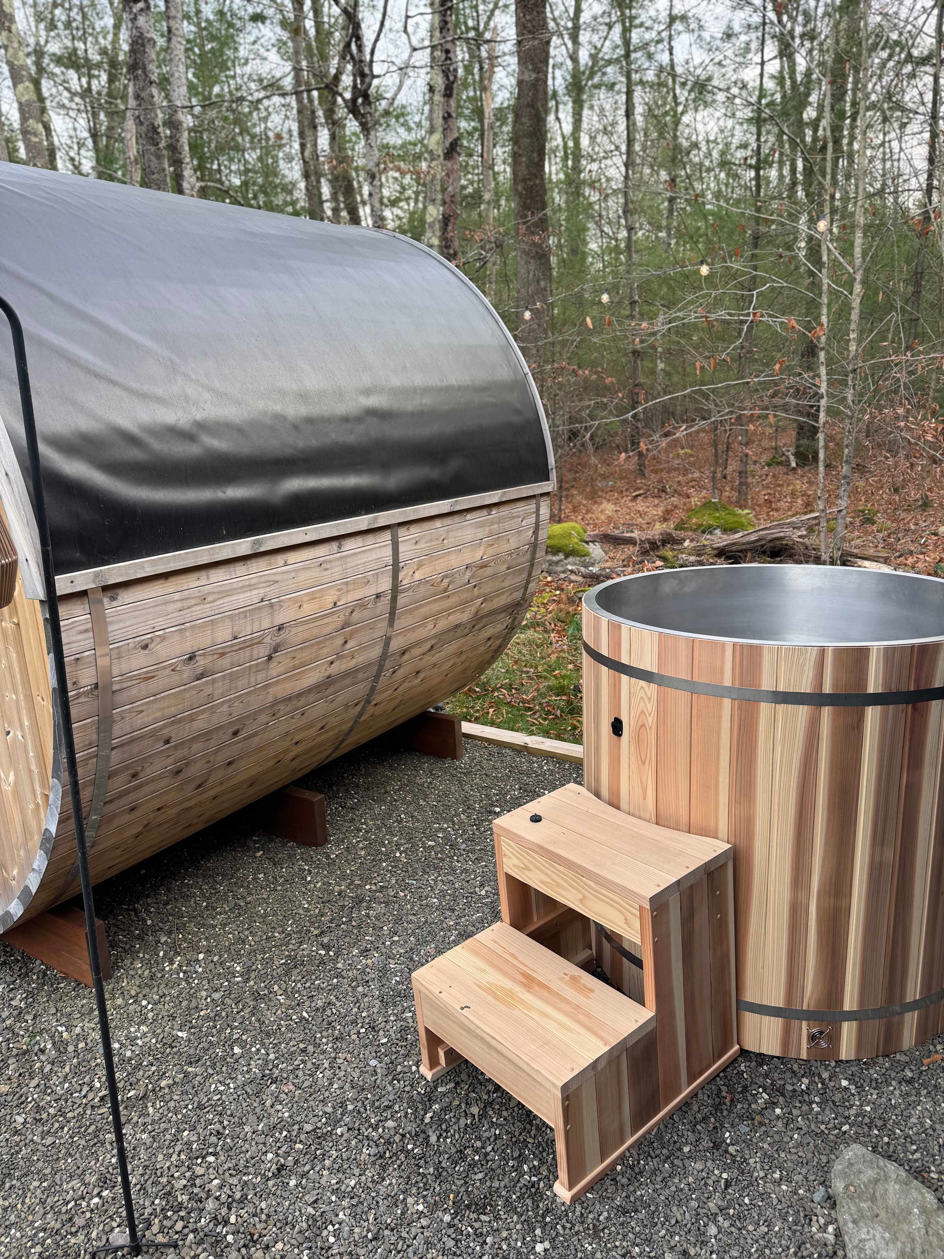 Exterior view of a wooden barrel sauna with a black roof, positioned next to a round wooden cold plunge tub with a metal interior, both set on a gravel surface.