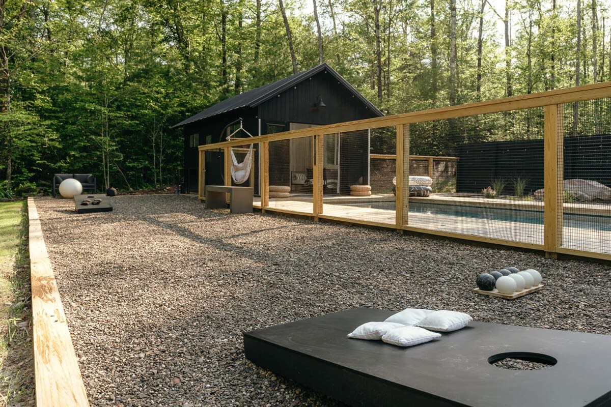 A gravel outdoor game area featuring a black cornhole board with white beanbags and a set of bocce balls, bordered by wooden planks. In the background, a black cabin and a swimming pool with a wooden deck are visible, surrounded by trees.
