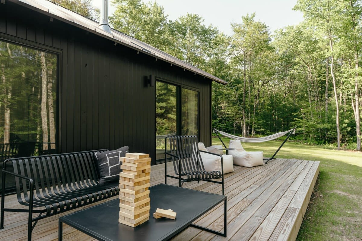 Wooden deck alongside the black-sided cabin, furnished with black metal outdoor seating, a coffee table with a Jenga game, and a white hammock in the grassy yard.
