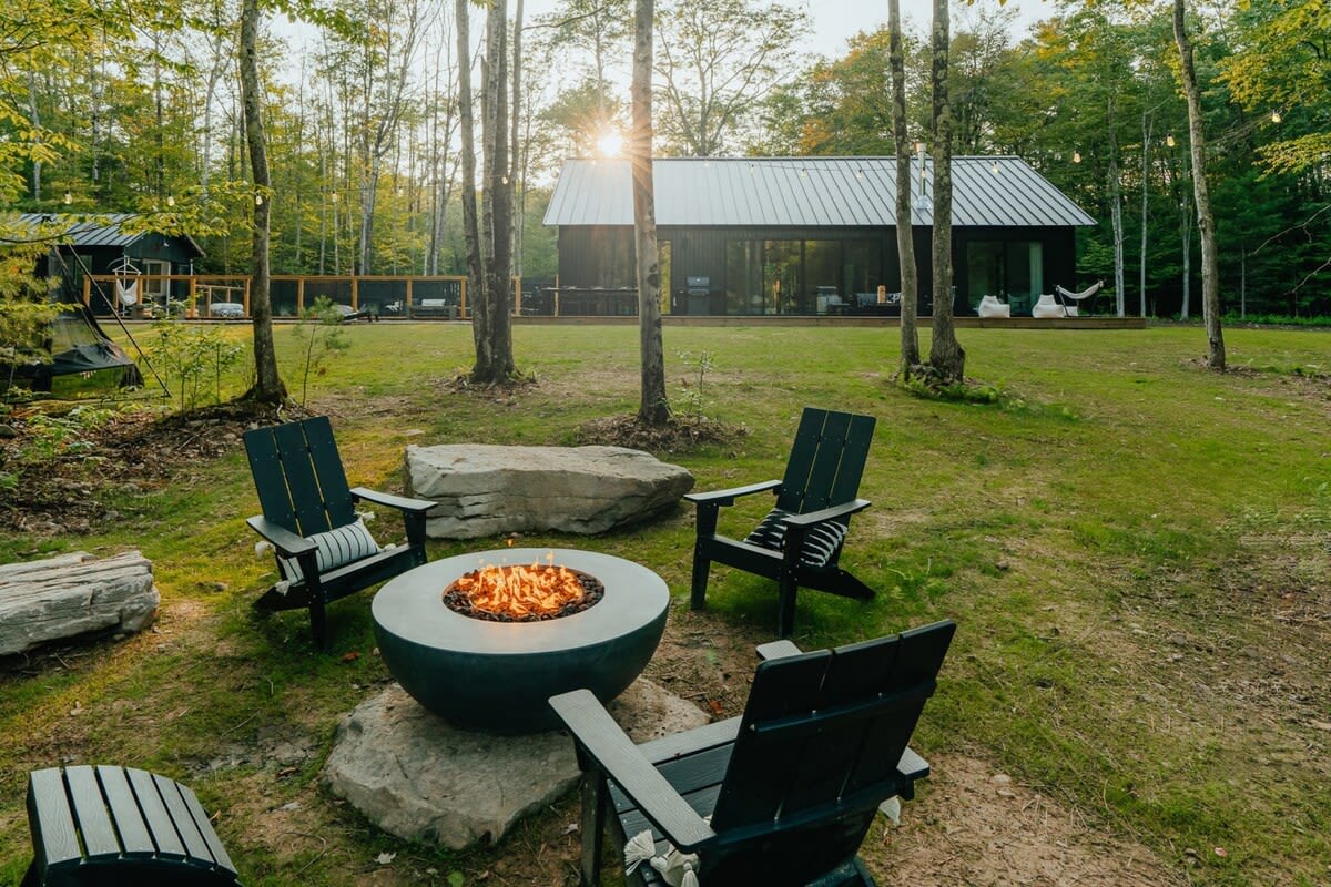 Outdoor fire pit area with a modern round fire bowl, surrounded by black Adirondack-style chairs and large rocks, set in a grassy clearing among tall trees with the cabin in the background.