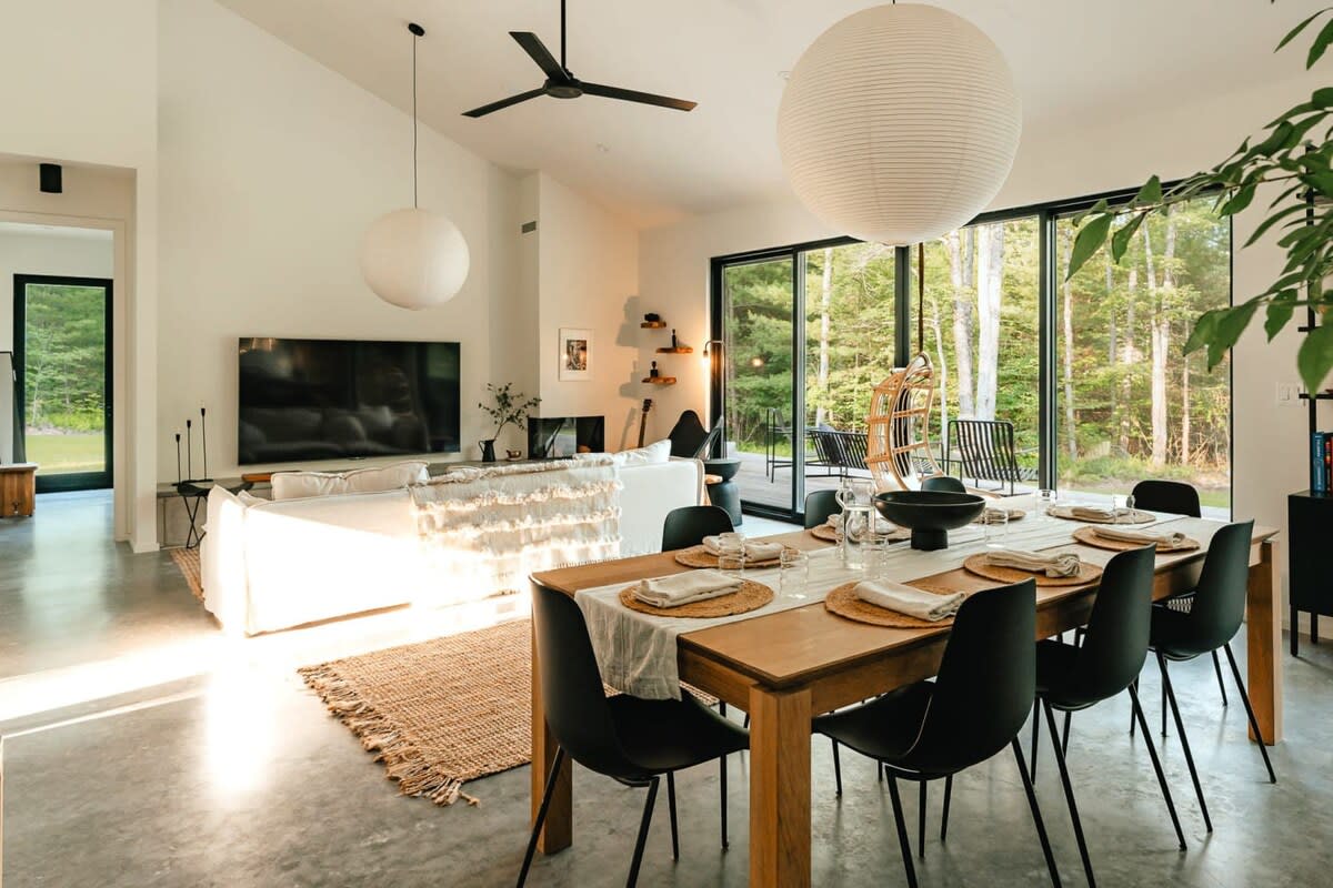 An open-concept living and dining area features a light wood dining table with black chairs, a white sectional sofa, and large sliding glass doors opening to a deck and forest. White walls, concrete floors, and modern light fixtures complete the space.