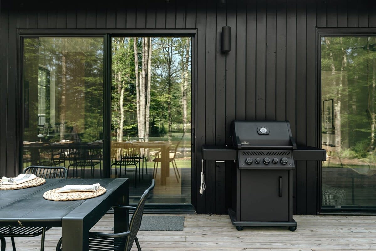 Part of the black-sided cabin exterior with large sliding glass doors, a black BBQ grill on a wooden deck, and reflections of the surrounding forest.