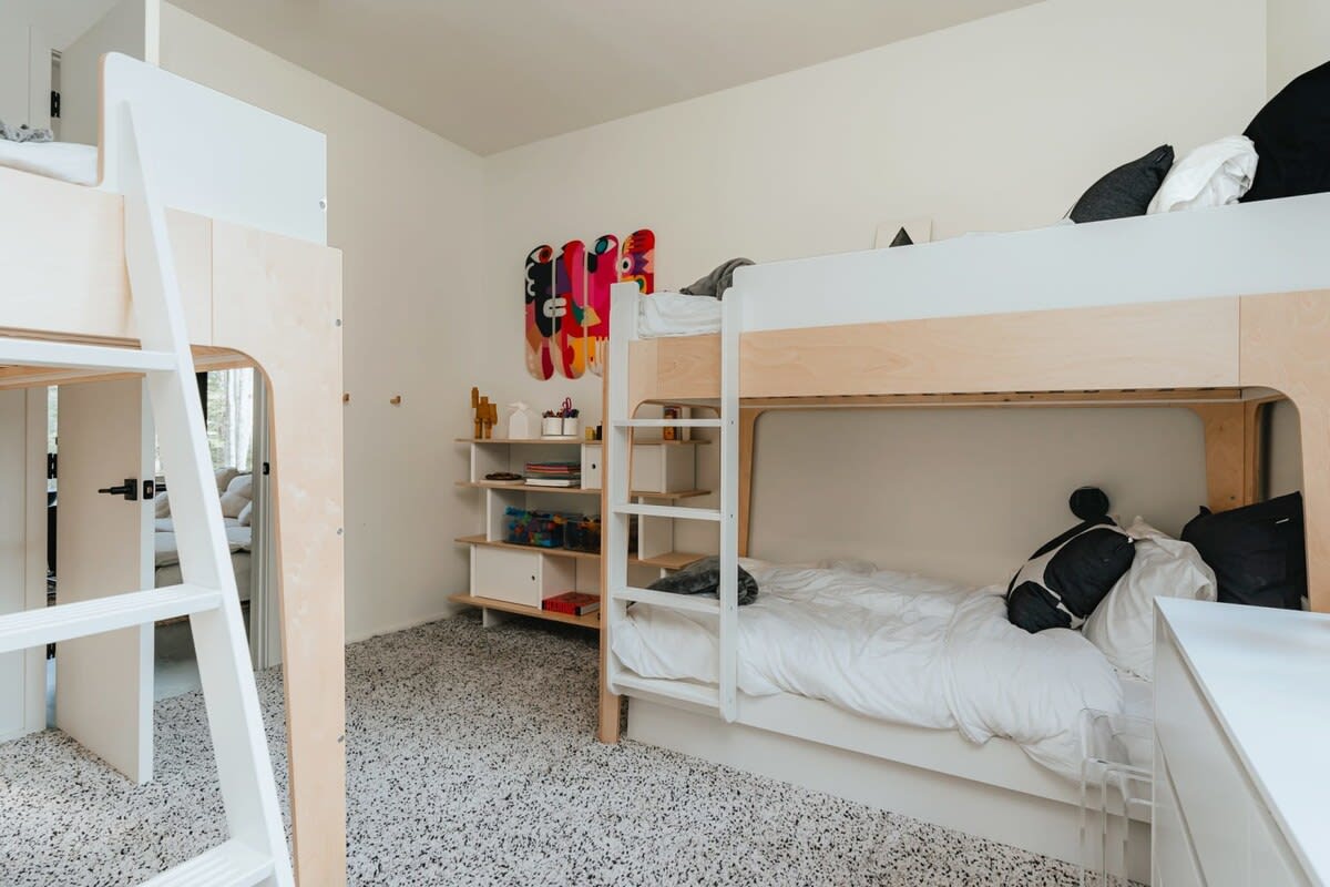 Another view of the kids' bedroom with light wood bunk beds, a white ladder, and open shelving with toys, set against a speckled carpet.