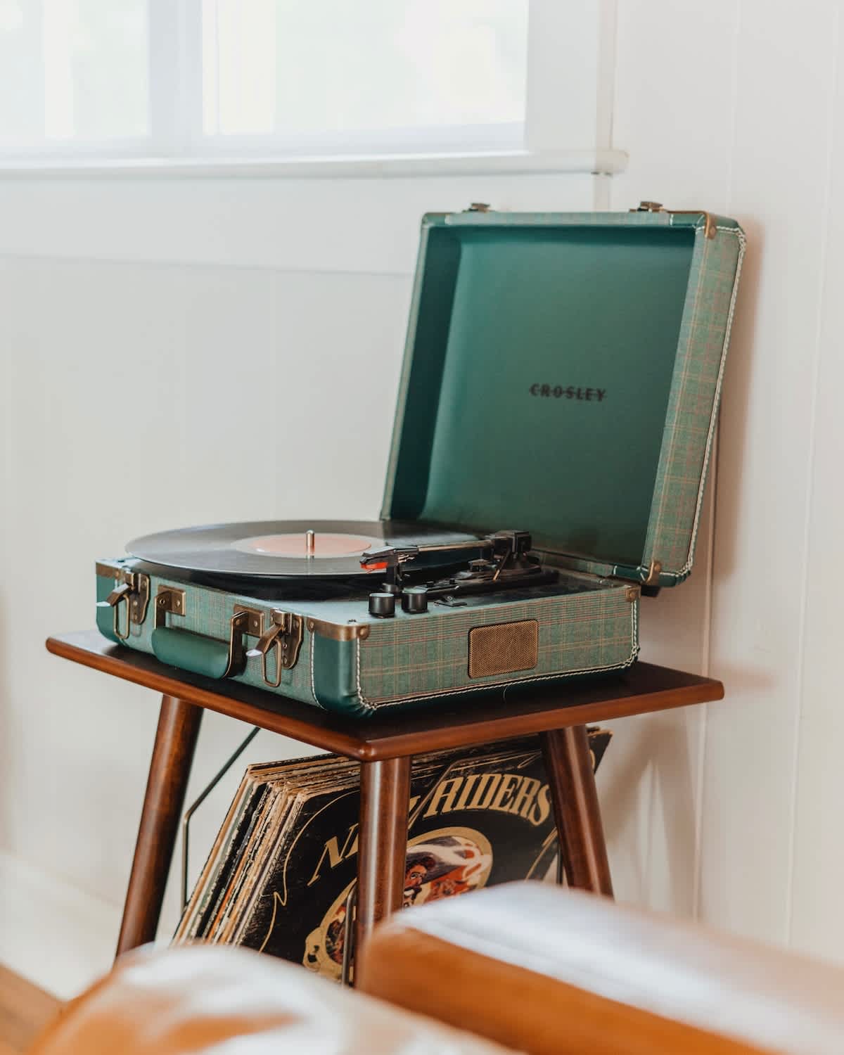 A vintage-style teal Crosley record player with a record spinning, placed on a mid-century modern wooden side table next to a stack of vinyl records, adding a retro touch to the living space.