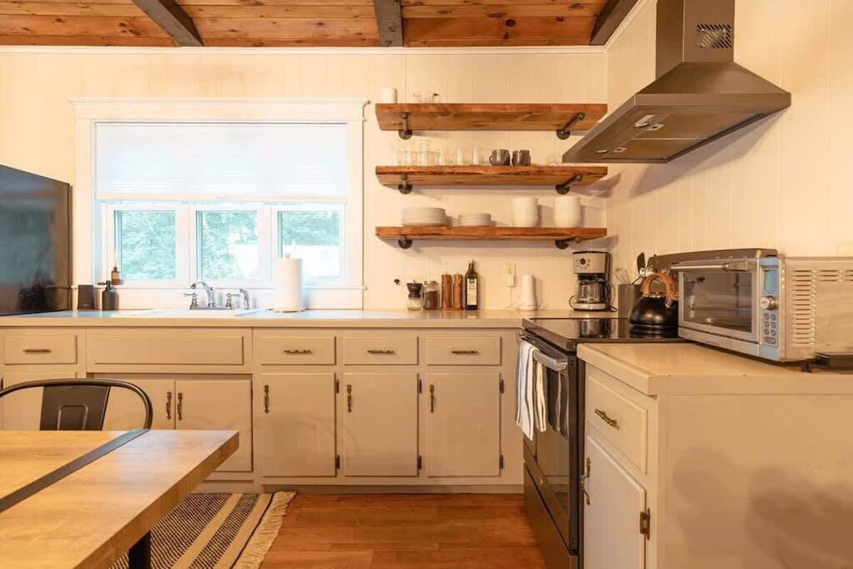 A functional kitchen area with light grey lower cabinets, a white countertop, a sink positioned under a window, and open wooden shelving above, equipped with a coffee maker and toaster oven.