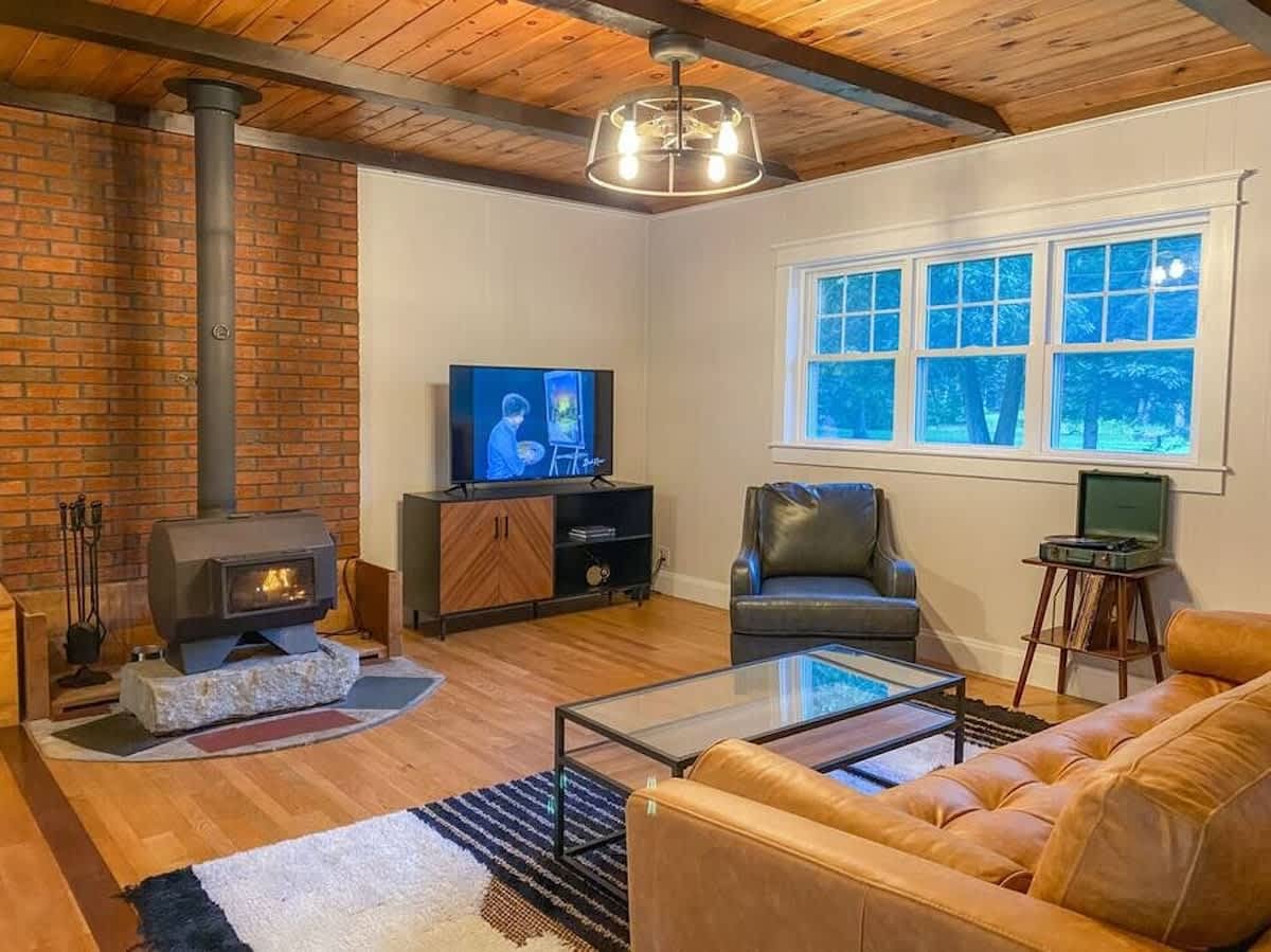 Cozy living room featuring a wood-burning stove on a stone hearth, a tan leather sofa, a dark leather armchair, and a flat-screen TV on a modern media console. Large windows overlook the outdoors.