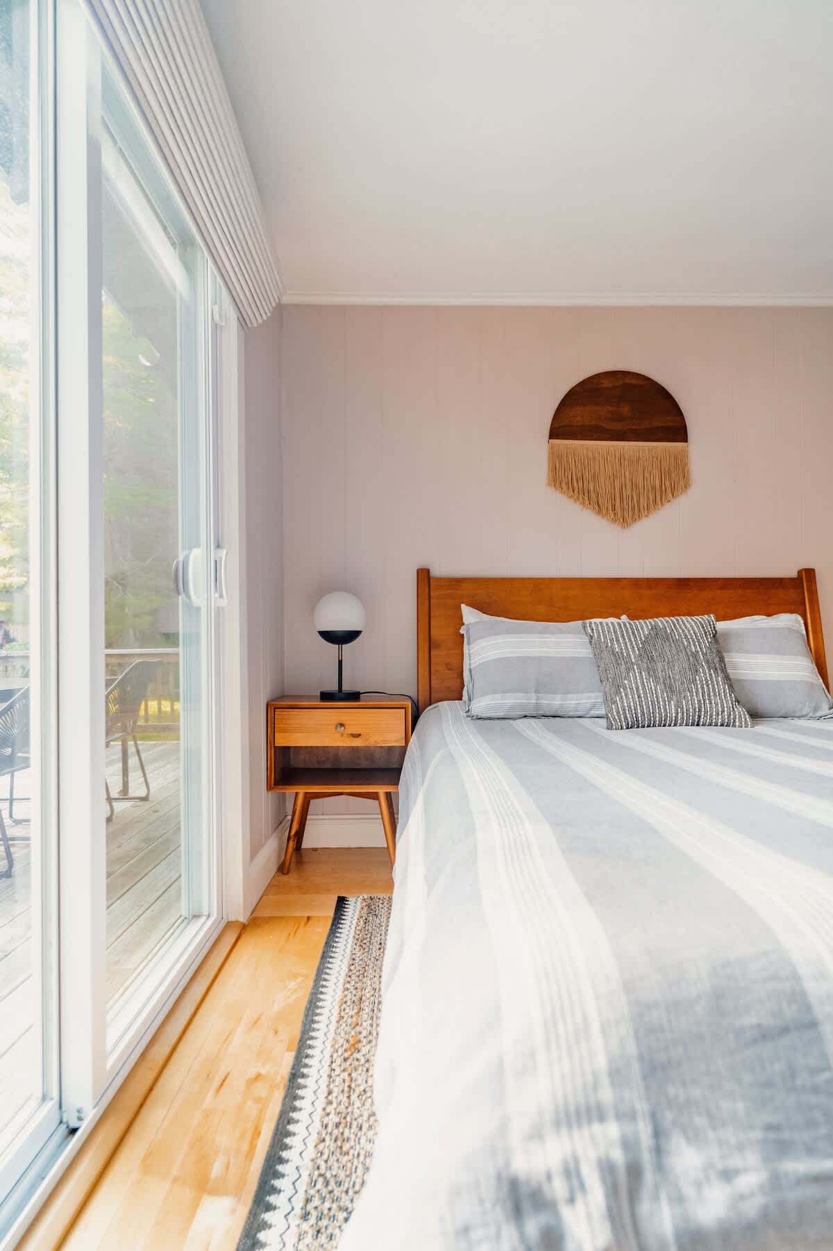 A bright bedroom features a wooden bed frame with striped bedding, a mid-century modern nightstand with a lamp, and a large sliding glass door offering access to an outdoor deck.