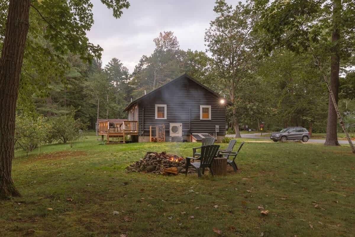 Exterior view of the dark-sided cabin with a wooden deck, surrounded by mature trees. A stone fire pit with Adirondack chairs is set in the grassy yard, ready for use.