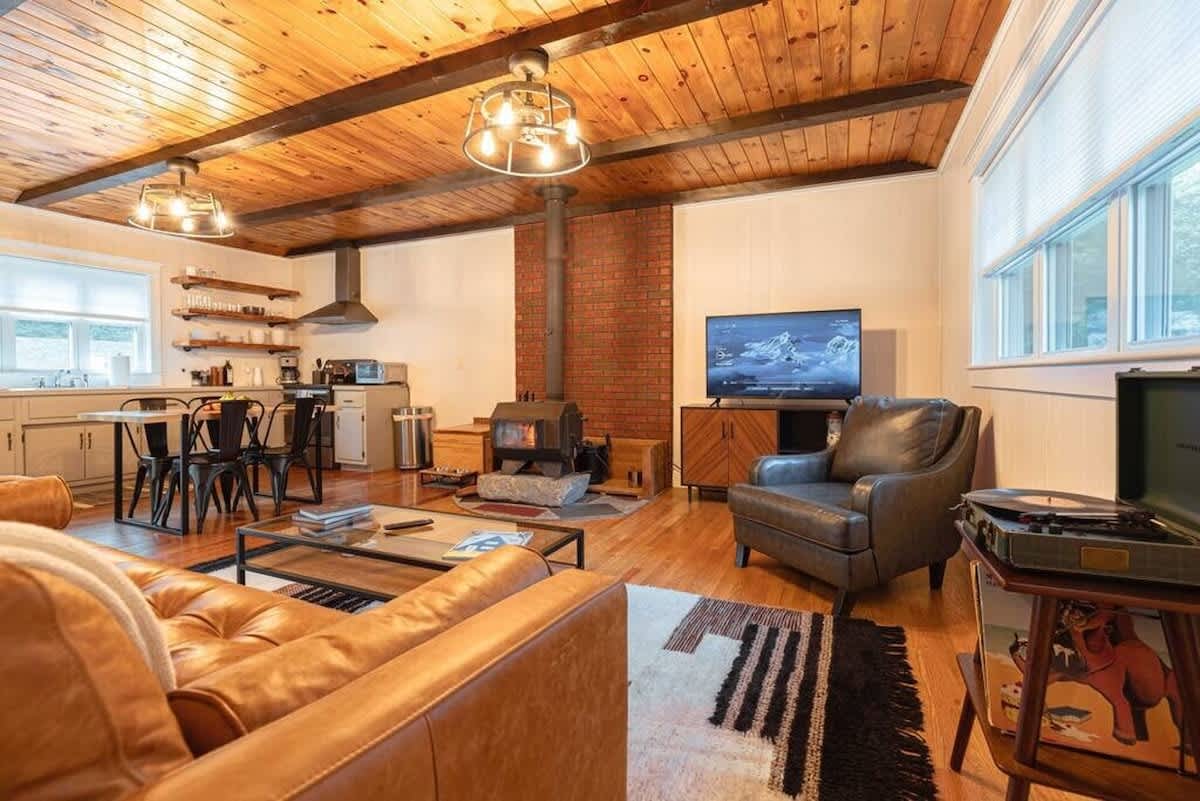 An inviting open-concept living space features a warm wooden plank ceiling with exposed beams, a cozy wood-burning stove set against a brick chimney, and a modern kitchen with light grey cabinets and open shelving.