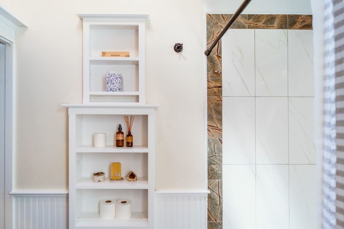 Bathroom with white built-in shelving holding toiletries and towels, adjacent to a tiled shower with a decorative stone border. White beadboard wainscoting lines the lower walls.