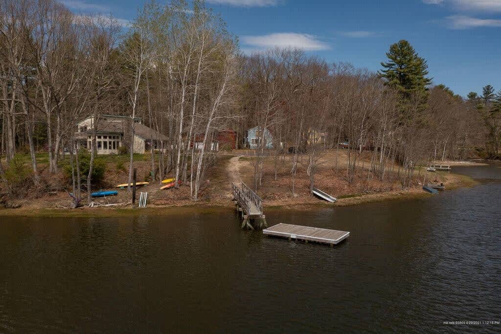 An aerial view of a calm body of water with a wooden dock extending into it, surrounded by a wooded shoreline with bare trees. A dirt path leads to distant houses, and several kayaks are visible on the shore.