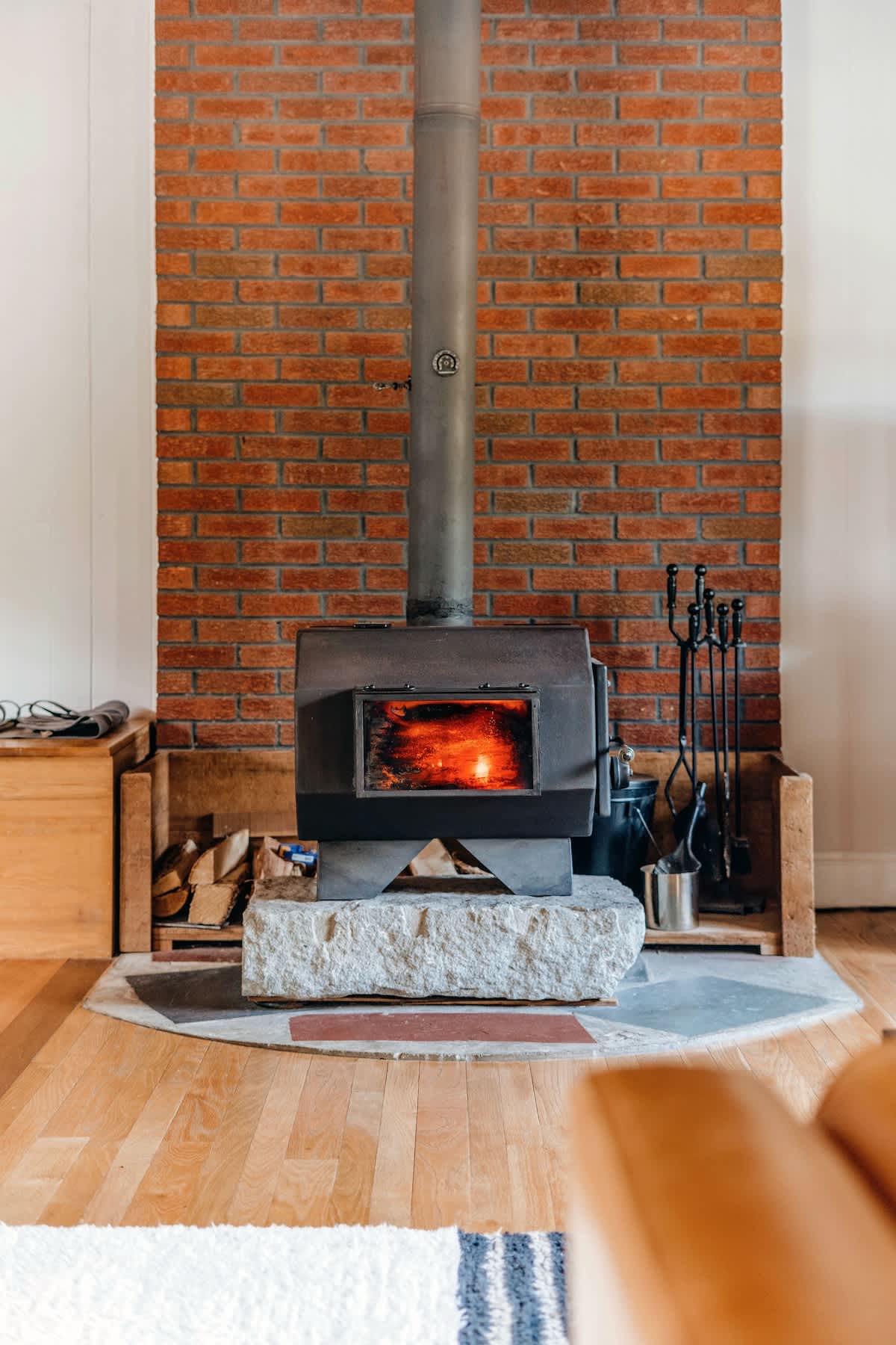 A close-up view of a black wood-burning stove with a visible fire, resting on a rough-hewn stone slab hearth, set against a rustic red brick chimney with stacked firewood on either side.