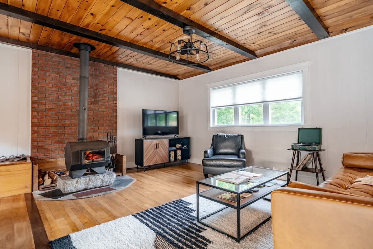 Inviting living room with a prominent black wood-burning stove set against a brick wall, complemented by a wood plank ceiling with dark beams. A tan leather sofa, black armchair, and TV console create a comfortable space.