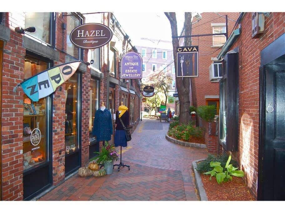 A quaint brick alleyway lined with boutique shops featuring display windows and hanging signs, with a tree providing shade in the middle. The brick path leads to a brighter area in the distance.