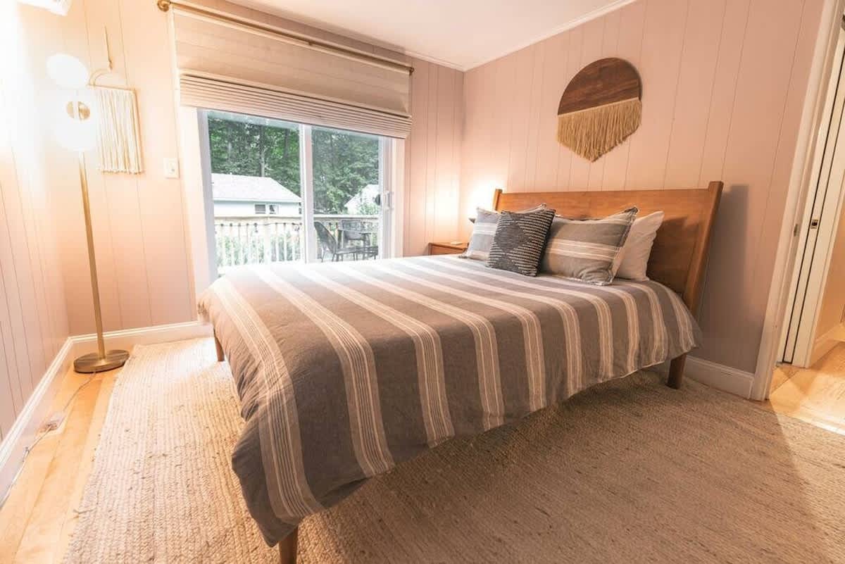 Serene bedroom with light pink vertical paneling and a wooden bed frame, dressed with striped bedding and decorative pillows. A large sliding glass door opens to an outdoor deck, providing natural light.