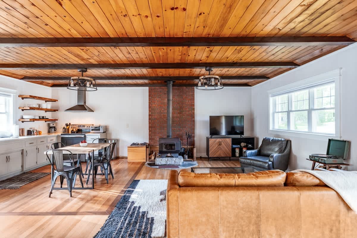 Expansive open-concept living and kitchen area showcasing a cohesive design with a wood plank ceiling, exposed beams, and light hardwood floors. The space includes a wood stove, comfortable seating, and a dining area.