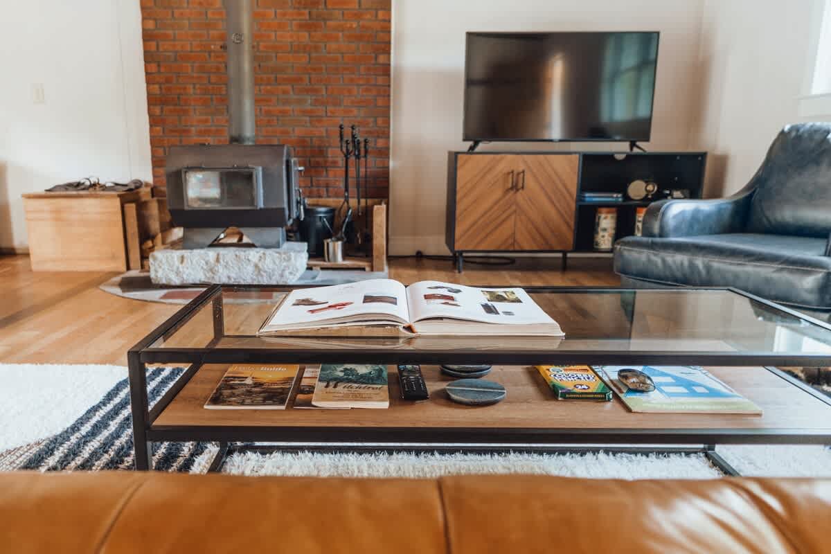A cozy living room view centered around a glass-top coffee table with books, showcasing a wood-burning stove, a flat-screen TV on a modern console, and a comfortable leather armchair.