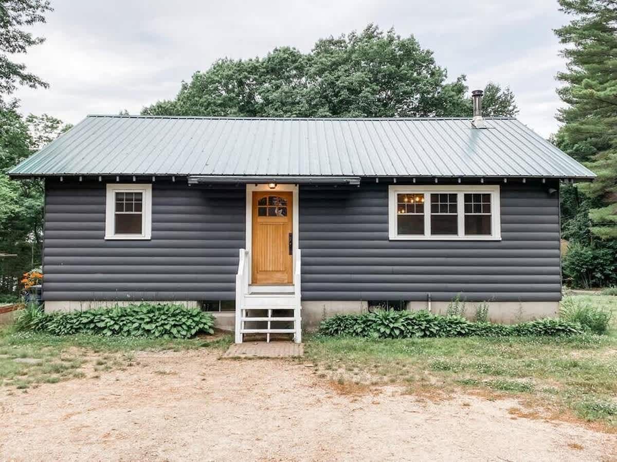 The front exterior of a dark gray, log-sided cabin with a light green metal roof, white window trim, and a natural wood front door. White steps lead up to the entrance, and green shrubs line the concrete foundation.