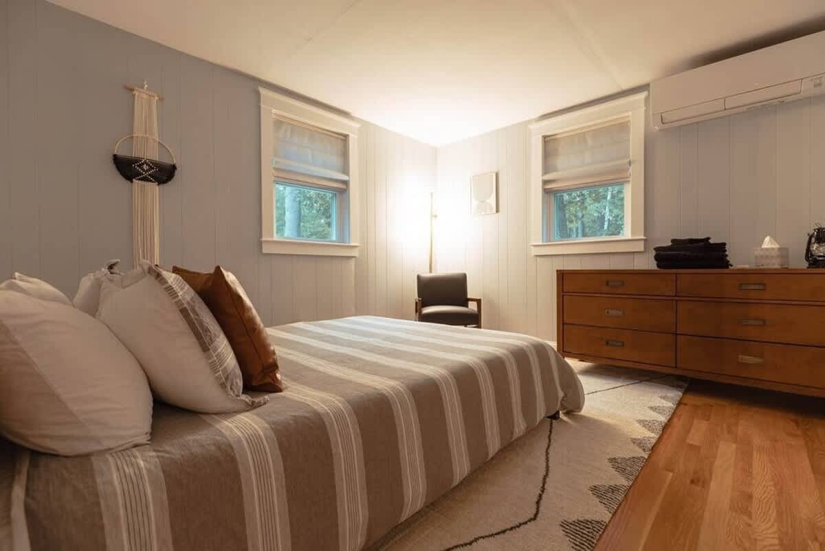 A cozy bedroom with light-colored vertical paneling and a striped bedspread, featuring a wooden dresser, a dark armchair, and two windows with room-darkening shades.