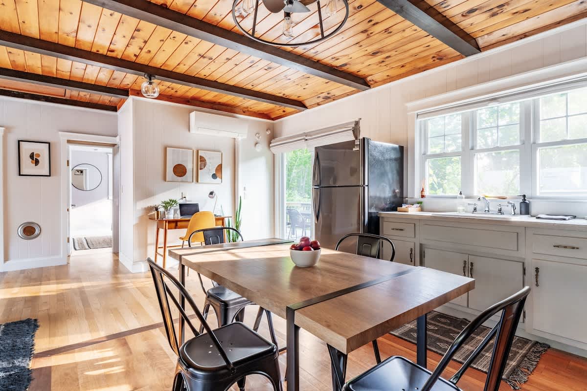 An open-concept space featuring a wooden dining table with black chairs, a modern kitchen with light grey cabinets and a black refrigerator, and a small office nook with a desk and chair.