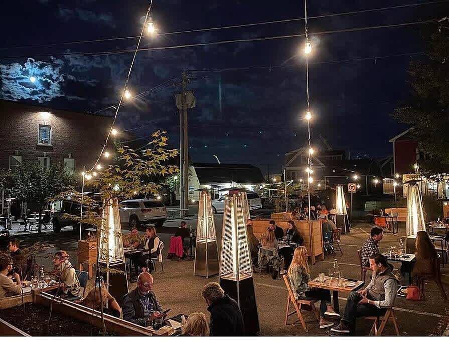 An inviting outdoor dining area at night, illuminated by string lights overhead and several tall patio heaters. Guests are seated at tables enjoying meals under a dark sky with a visible moon.