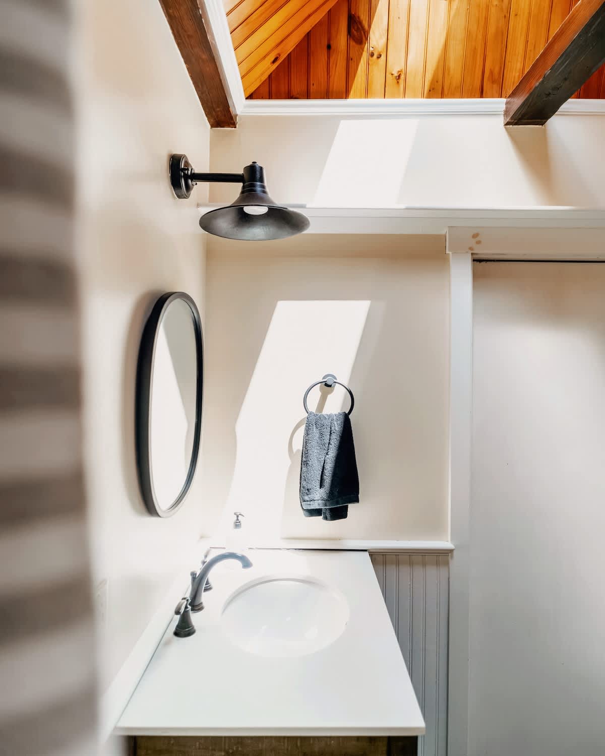 Bright bathroom vanity with a white sink, dark faucet, and a round black mirror. A wall-mounted light fixture and a towel ring are visible, with natural light streaming in from above.