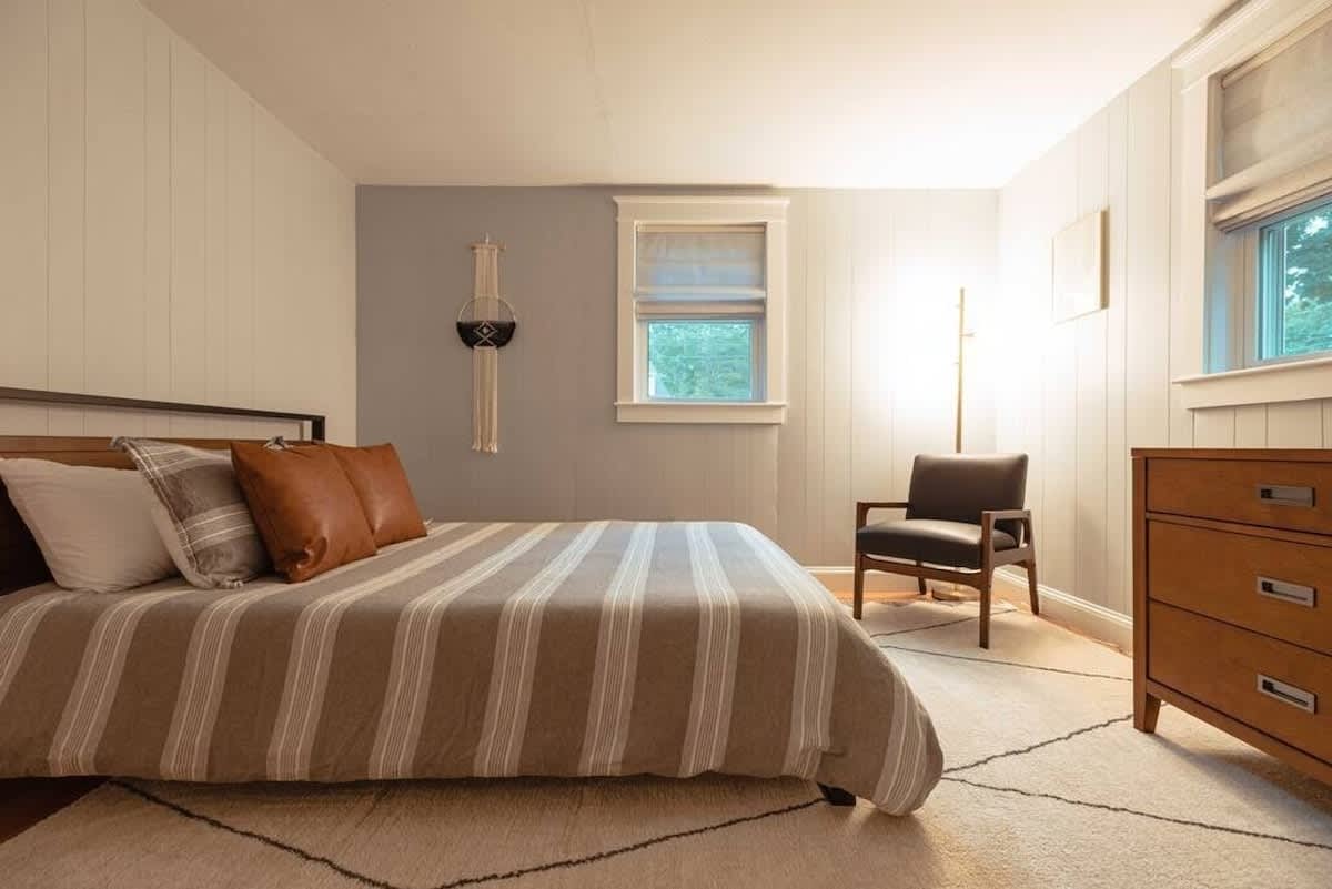 Another perspective of the bedroom with light paneling, showing a bed with striped bedding and brown pillows, a wooden dresser, a floor lamp, and a dark armchair, all contributing to a comfortable ambiance.