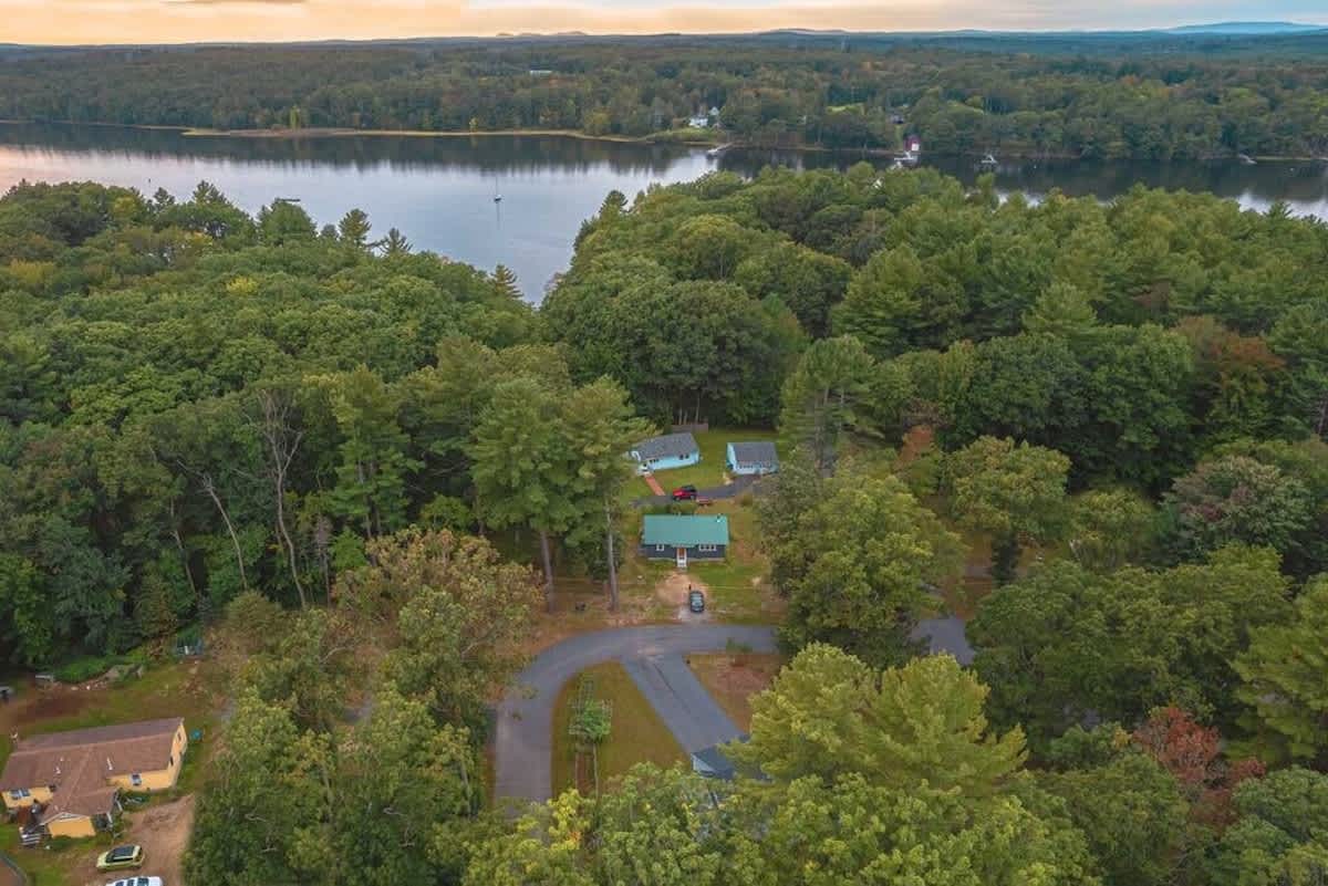 Aerial view of the cabin nestled among lush green trees, with a winding road leading to it and a large body of water (lake or river) in the background under a cloudy sky.