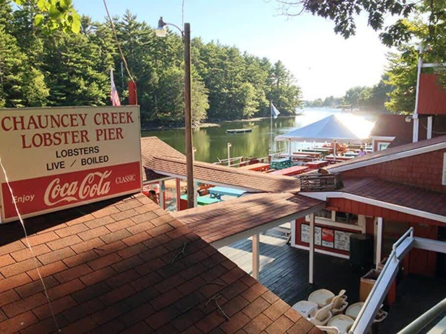An elevated view of "Chauncey Creek Lobster Pier," a red-roofed restaurant with outdoor seating overlooking a calm body of water surrounded by dense green trees. A large sign advertises live and boiled lobsters.