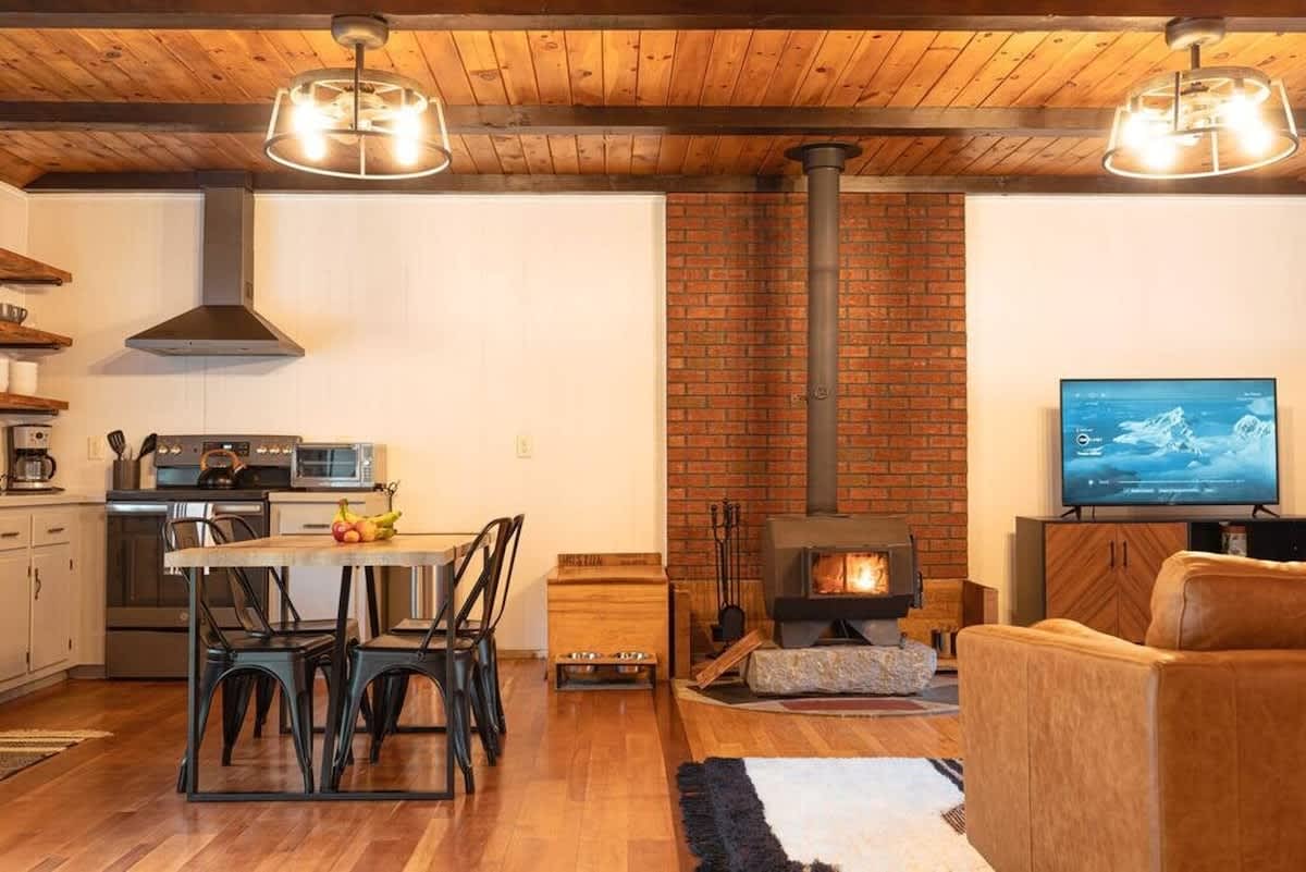 Open-concept living space with a wood-burning stove on a brick accent wall, a dining table with black chairs, and a modern kitchen with stainless steel appliances. Wooden beamed ceiling adds rustic charm.