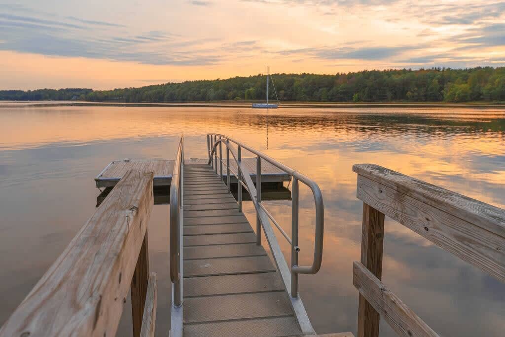 A sturdy metal and wood dock extends into a calm body of water, reflecting the warm hues of a sunset sky. A sailboat is visible in the distance, surrounded by forested shores.