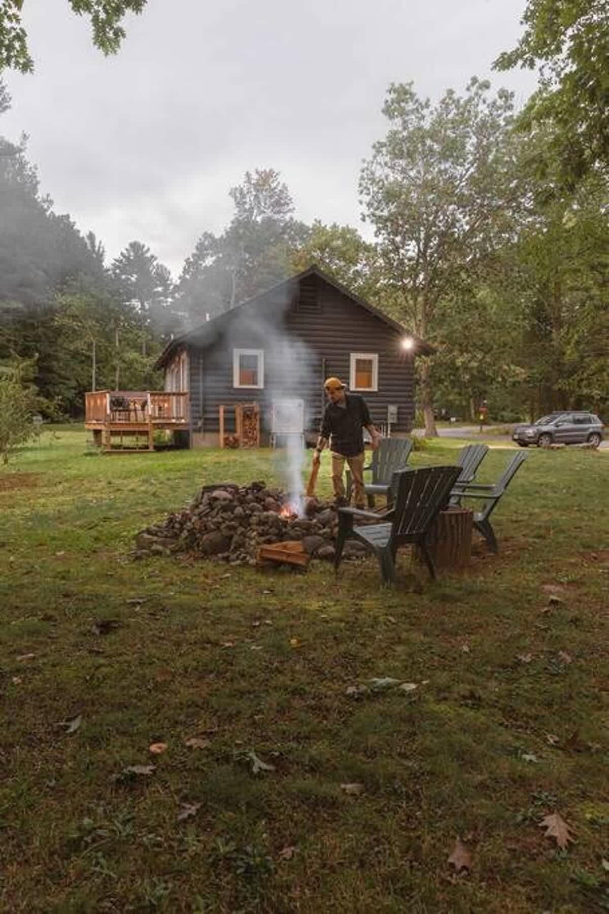 A person tends to a smoking fire pit surrounded by Adirondack chairs in the grassy yard, with the dark-sided cabin and its wooden deck visible in the background under an overcast sky.