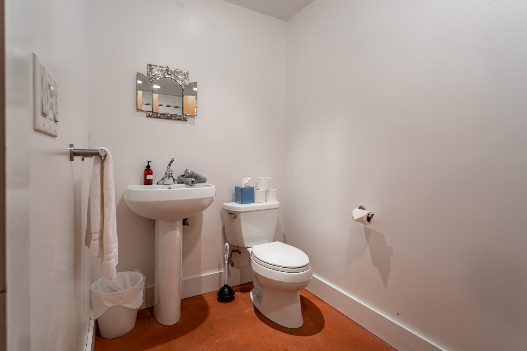 Compact bathroom featuring a white pedestal sink, a toilet, and a decorative mirror above the sink, with a warm-toned floor.