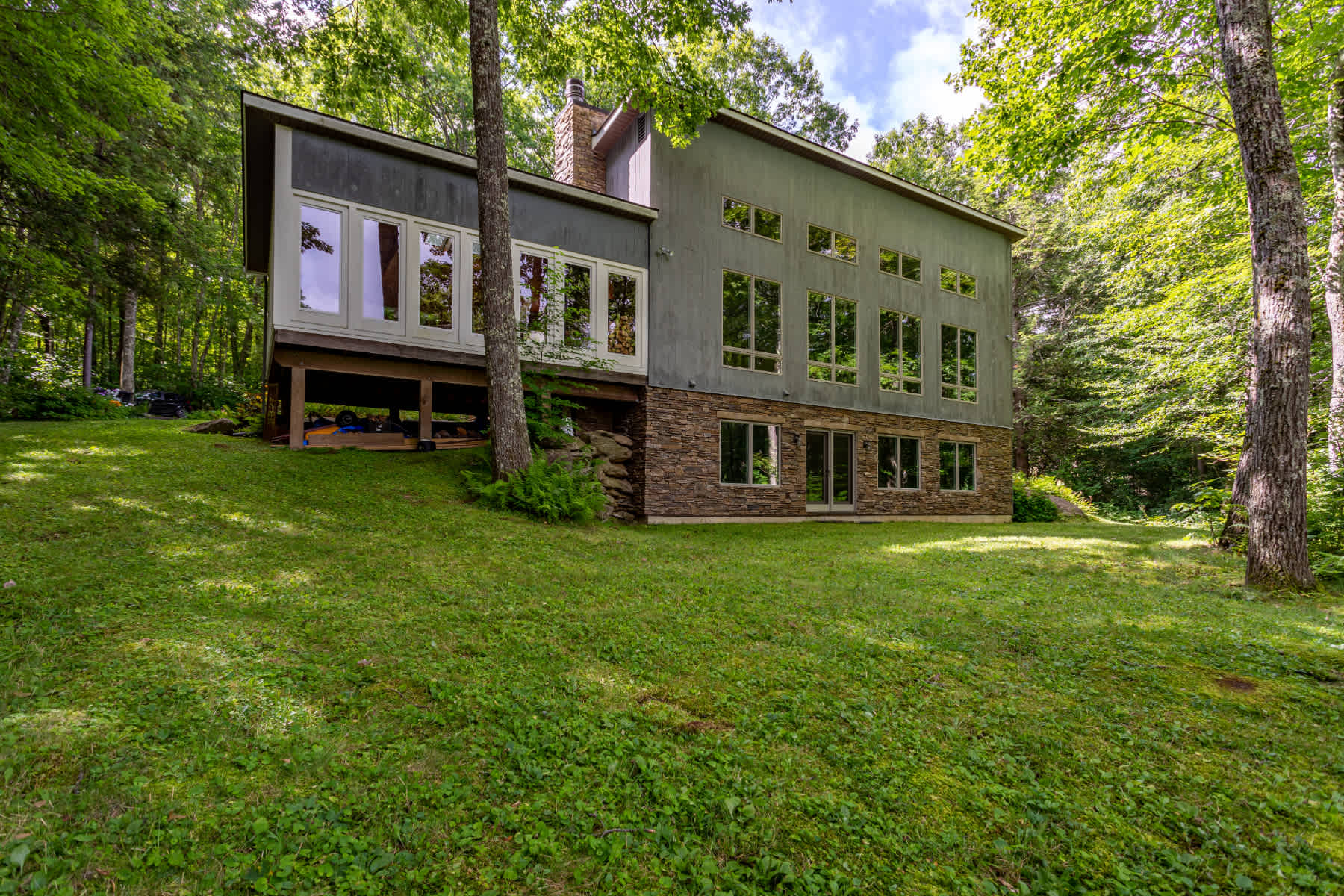 Modern multi-level house with a grey facade, large windows, and a stone-clad lower level, nestled on a grassy slope amidst lush green trees under a partly cloudy sky.