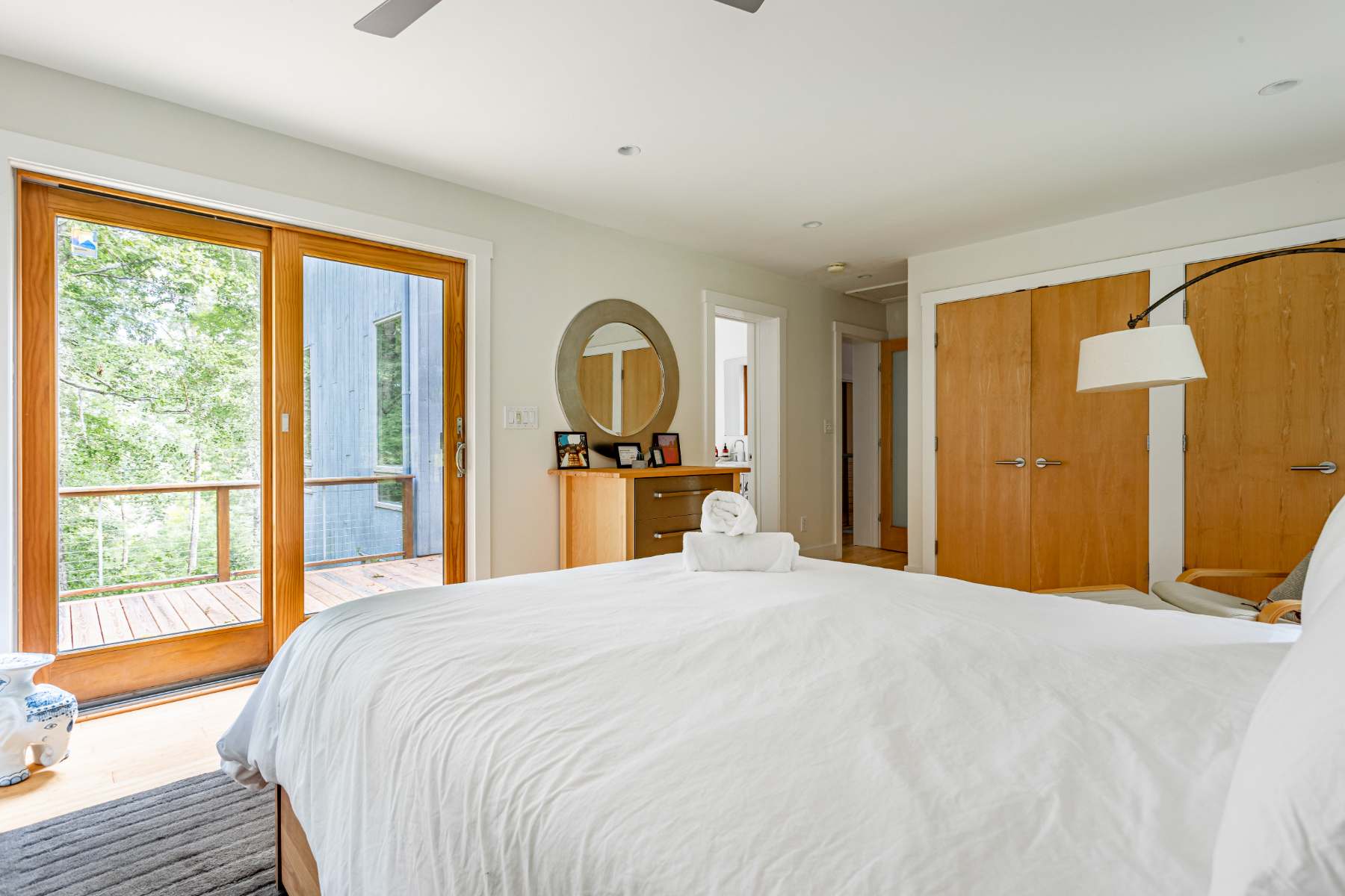 View of a bright bedroom with a white bed, a sliding glass door opening to a deck, and light wood closet doors, complemented by a dresser with a round mirror.