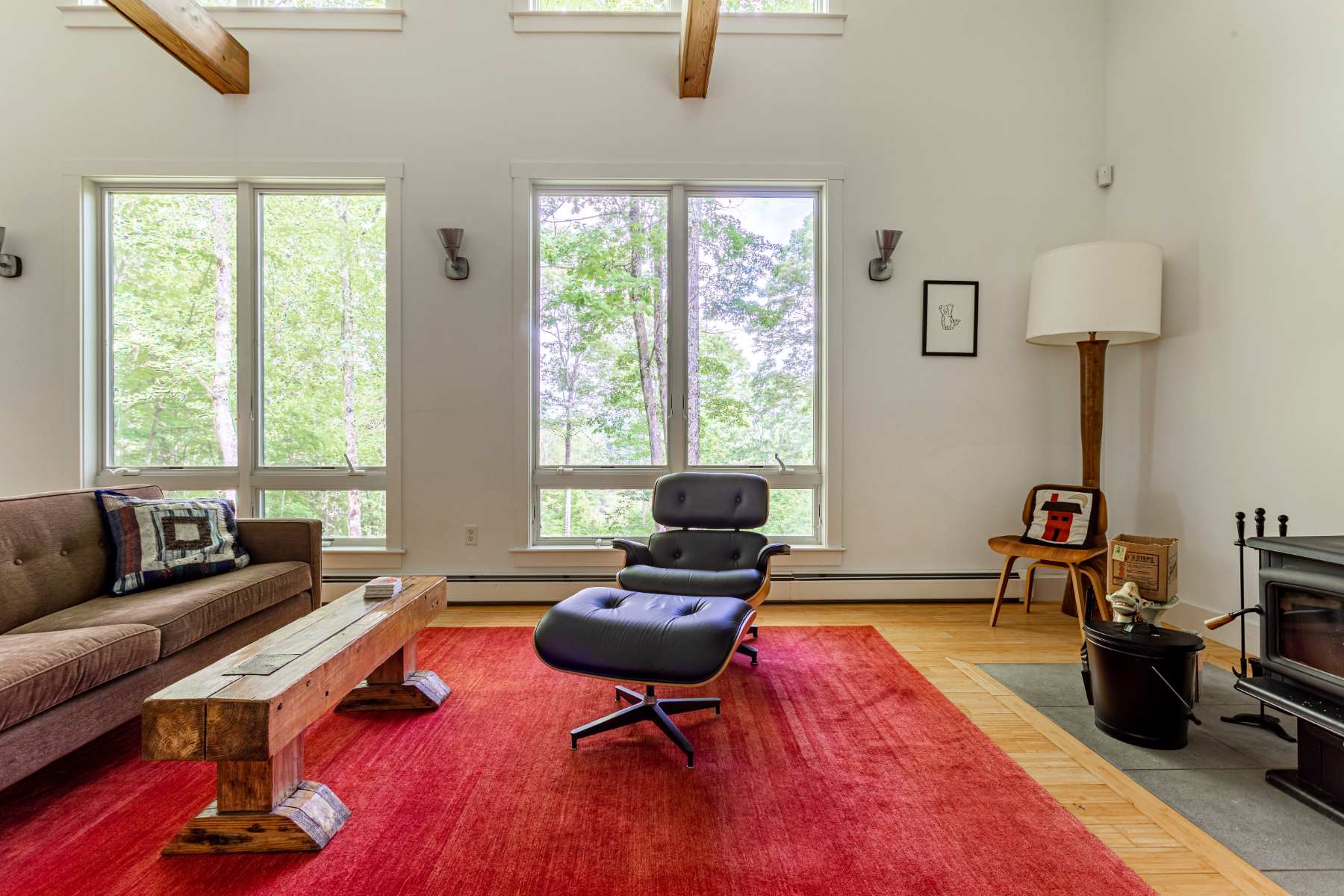 A spacious living room features a brown sofa, a black leather armchair with an ottoman, and a rustic wooden coffee table on a vibrant red area rug. Large windows offer views of the surrounding trees, complemented by high white walls and exposed wooden beams.