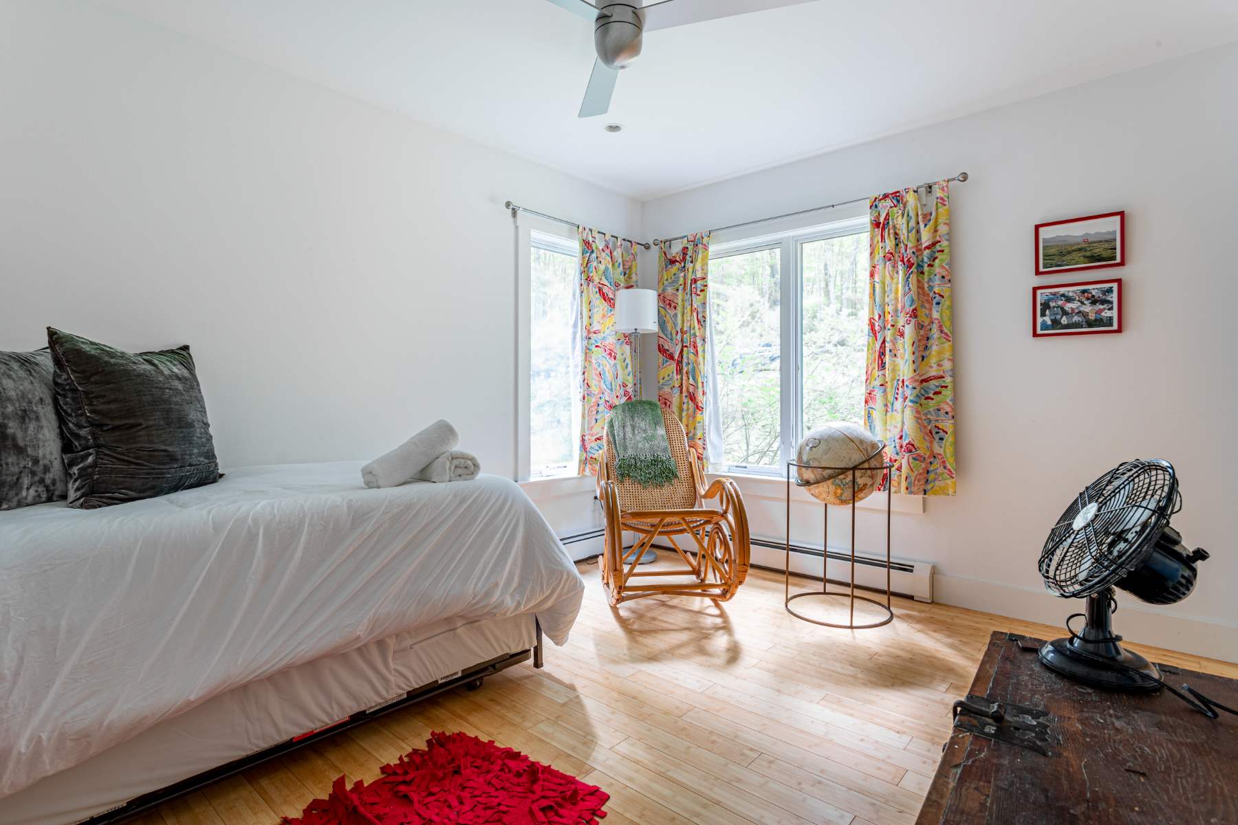 A bright bedroom with light hardwood floors features a single bed with white linens and dark grey pillows. A wicker rocking chair and a globe on a stand are placed near large windows with colorful floral curtains, offering views of the outdoors.