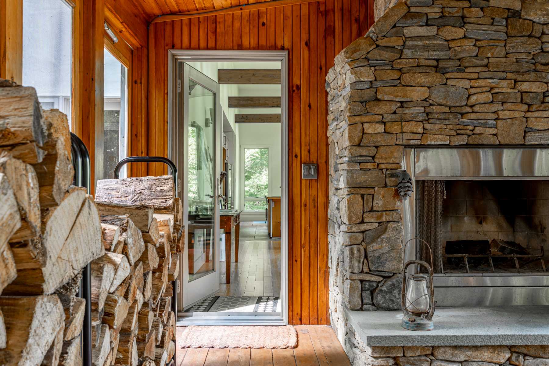 A close-up of a rustic stone fireplace with a metal lantern on the hearth, flanked by neatly stacked firewood. A glass-paned door opens to an interior hallway with light wood floors and white walls, offering a glimpse into the main living space.